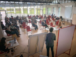 Auf dem Bild sieht man eine große Anzahl an Menschen, die auf roten Stühlen sitzen. Sie diskutieren miteinander und im Vordergrund sieht man eine Person zwischen zwei Flipcharts stehen